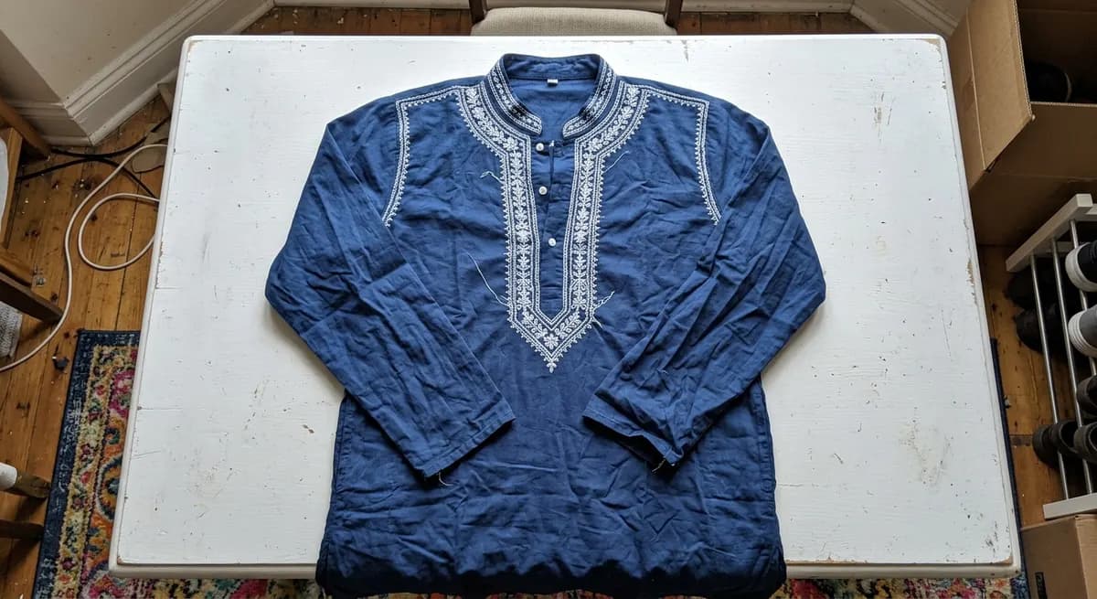 Amateur flat-lay photo of a blue cotton kurta taken with a phone camera on a plain surface — typical before image uploaded to ShotRoom for AI virtual photoshoot transformation